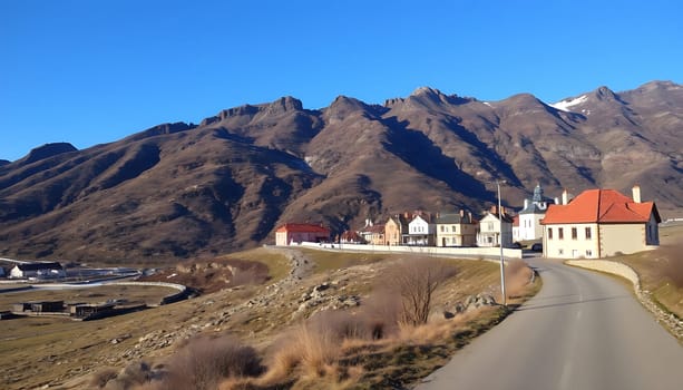 A scenic image of a winding road leading up to a small village nestled against a backdrop of majestic mountains. The houses, with their colorful facades, add a touch of charm to the rural landscape. The clear blue sky and the sun-drenched slopes create a sense of tranquility and adventure. This photo is ideal for use in travel brochures, websites, and social media posts promoting mountain destinations. It captures the essence of rural life and the beauty of nature.
