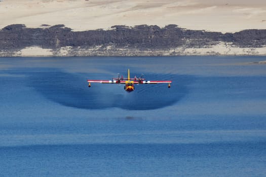 A firefighting plane collects water from a calm lake against a backdrop of sandy shores,