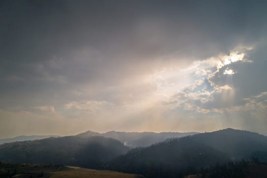 A breathtaking view of sun rays piercing through dramatic clouds over lush, misty mountains at dusk. A serene and majestic scenery evoking tranquility and awe.