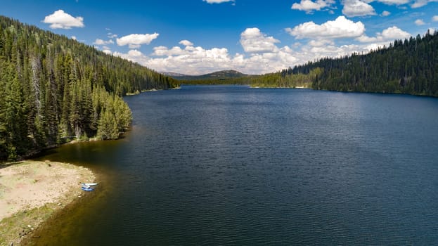 A serene mountain lake with calm water and lush pine forests under a vibrant blue sky with fluffy clouds, offering a peaceful natural retreat.