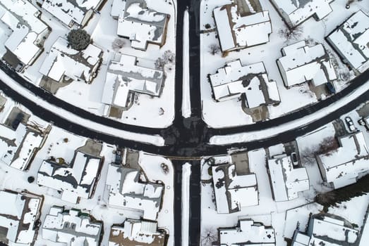 Aerial shot featuring a snow-covered suburban neighborhood during winter. The intersecting streets and snow-laden rooftops create a serene and symmetrical winter landscape.