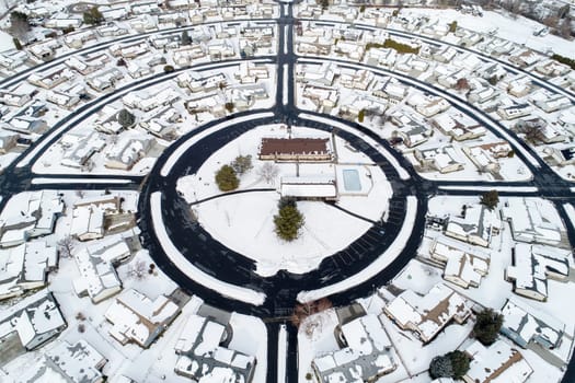 High-angle aerial photograph of a circular suburban neighborhood blanketed in snow, showcasing smooth roundabouts and rooftops. A peaceful winter scene captured from above.