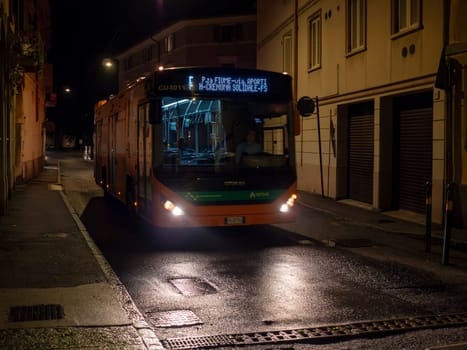 Cremona, Lombardy, Italy October 15th 2024 An electric bus is driving down a narrow, empty street at night, illuminating the wet asphalt with its headlights