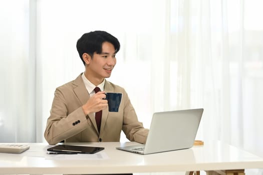 Young businessman drinking coffee and working with laptop in bright office.