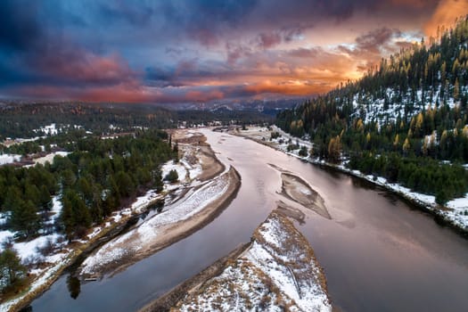A stunning aerial view of a snow-covered river cutting through a forested landscape during a vibrant sunset. Captures the tranquility and beauty of nature in winter.