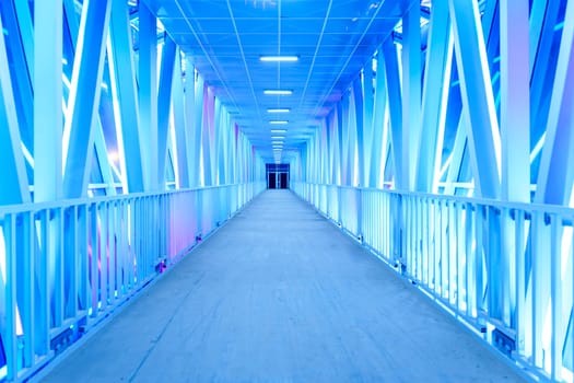A futuristic illuminated walkway with a symmetrical design and glowing blue lights creates a modern, minimalist atmosphere. The empty corridor stretches into the distance with no people in sight