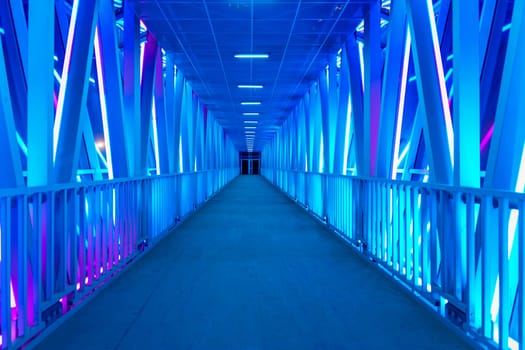A futuristic illuminated walkway with a symmetrical design and glowing blue lights creates a modern, minimalist atmosphere. The empty corridor stretches into the distance with no people in sight
