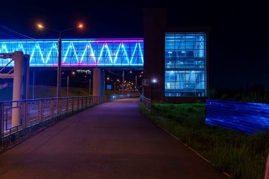 An illuminated pedestrian bridge with vibrant blue and purple lights connects to a modern glass building at night. The scene is peaceful and urban, with no people in sight