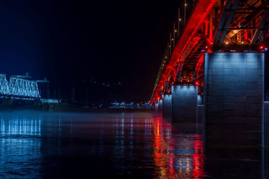 Red and blue illuminated bridges stretch over a river at night, with mist hovering over the water and reflections shimmering below. The serene urban scene captures the contrasting colors