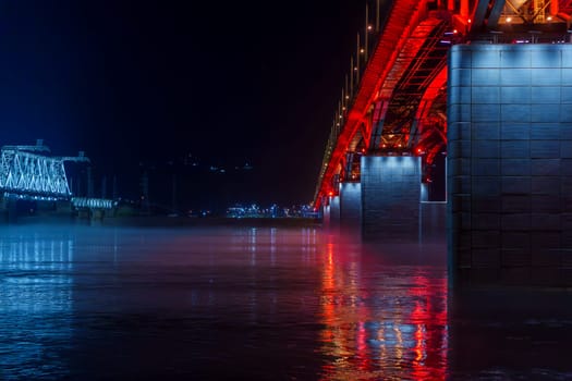 Red and blue illuminated bridges stretch over a river at night, with mist hovering over the water and reflections shimmering below. The serene urban scene captures the contrasting colors