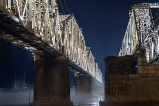A steel bridge illuminated at night spans over a calm river with mist hovering above the water. The serene scene highlights the structure and lighting with no people present