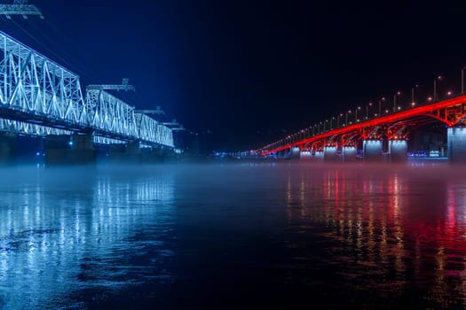 Two illuminated bridges, one blue and one red, stretch over a river at night. A layer of mist hovers over the water, reflecting the lights from the bridges