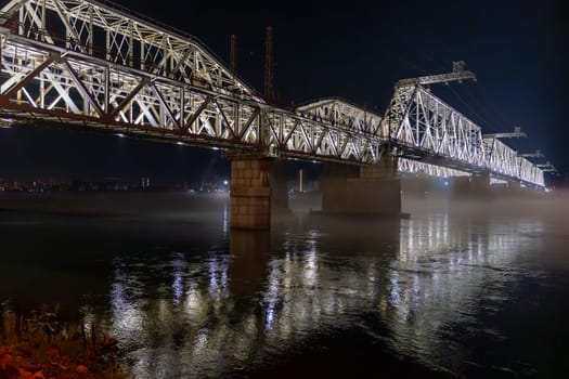 An illuminated steel bridge stretches over a calm river at night, with mist hovering above the water and reflections of the lights. The peaceful scene is captured with no people present