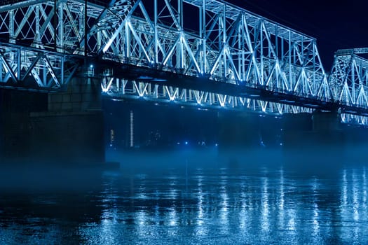 A blue illuminated steel bridge spans over a calm river at night, with mist hovering above the water and reflections shimmering below. The peaceful and serene urban scene shows no people present