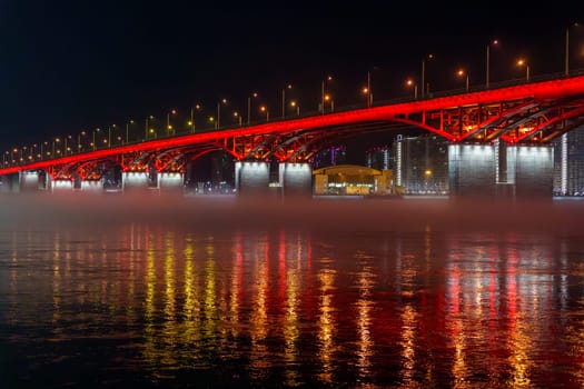 A red illuminated bridge stretches over a calm river at night, with mist floating above the water and colorful reflections. The serene urban scene captures the structure and lighting