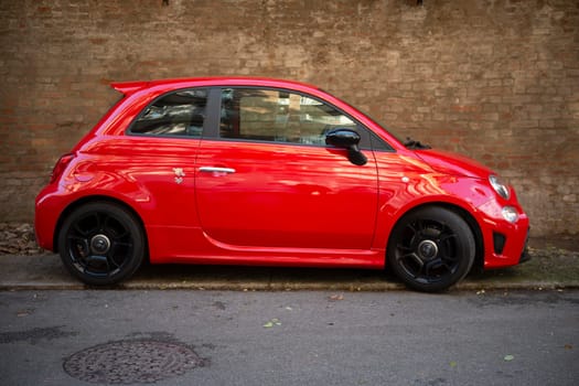 Cremona, Lombardy, Italy October 15th 2024 A vibrant red fiat 500 abarth tuned parked gracefully on a bustling city street against a rustic brick wall, embodying luxury and speed in its compact design