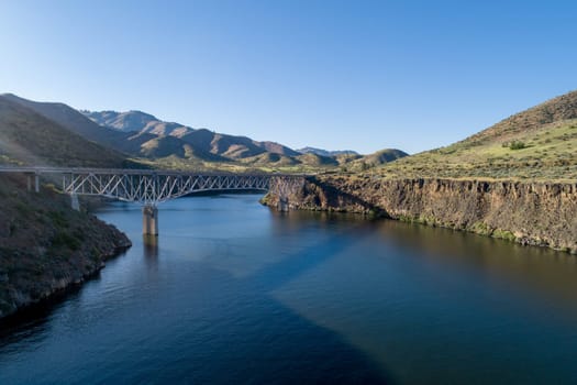 Aerial view of a steel bridge crossing a tranquil river, framed by lush green hills under a clear blue sky, capturing the essence of peaceful nature and engineering beauty.