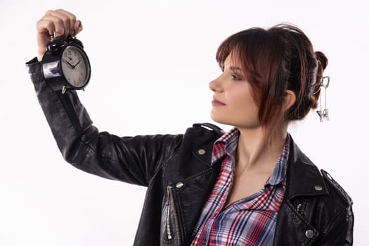 A young woman in a leather jacket and plaid shirt thoughtfully examines an alarm clock, reflecting on time