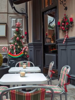 The image depicts a festive outdoor dining area decorated for Christmas. The centerpiece is a Christmas tree adorned with red and gold ornaments and a star on top, surrounded by candy canes. The tree is placed on a black table with a white tablecloth. The table is set with two candles and two glasses. The dining area is furnished with gray chairs with red and green plaid cushions, and a black awning provides shade. A black door with a window is visible in the background.