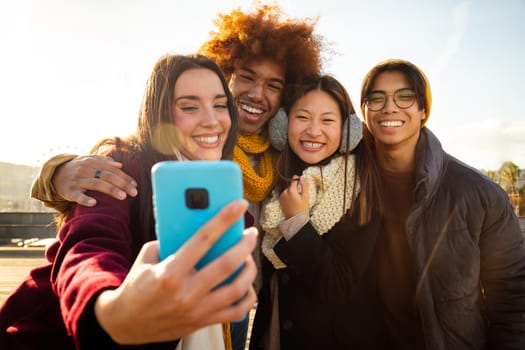 Young caucasian female taking selfie with group of multiracial friends outdoors in winter sunny day using mobile phone. Technology and lifestyle concept.