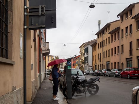 Cremona, Lombardy, Italy October 15th 2024 Woman using parking meter on rainy italian street, holding red umbrella and protecting herself from bad weather
