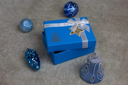 New Year gift in blue decorated box with bow with ajar lid next to New Year toys for Christmas tree on light background, concept for new year gift in packaging