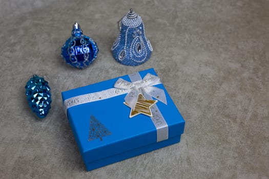 Gift box for Christmas, New Year gift in beautiful package lies on light background in front of New Year toys for Christmas tree, cute box for winter holiday gift