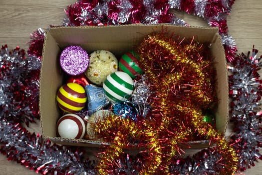 Tinsel and New Year toys for Christmas tree in cardboard box on background of floor, box for storing Christmas decorations at home, preparing to decorate house for winter holidays