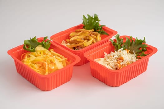 Three red plastic containers holding an assortment of salads and fries with garnishes, for takeaway or ready-to-eat meals.
