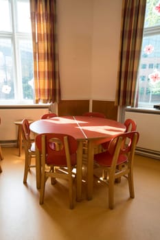 A bright and colorful classroom corner featuring a round table with wooden chairs, illuminated by sunlight. Decorated walls and plaid curtains create a welcoming learning environment.