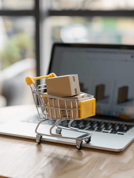 A metal shopping cart filled with cardboard boxes sits on a laptop, illustrating the connection between digital ordering and the physical fulfillment of ecommerce purchases