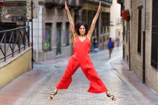 A woman dressed in a bright, vibrant red jumpsuit joyfully leaps and jumps energetically on a picturesque city street, radiating pure happiness, positivity, and exuberant energy for all to see