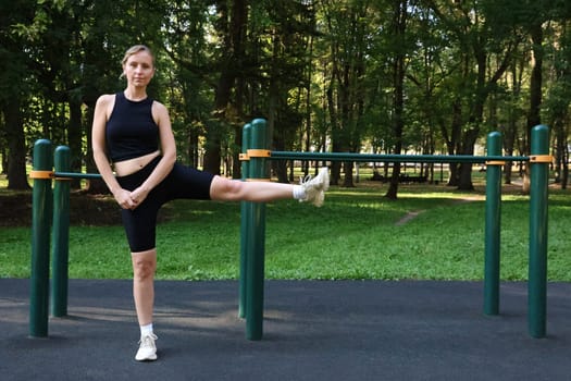 Slender woman in black sports uniform doing physical exercise against the background of sports ground. Outdoor Sports.