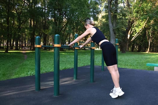 Workout, woman doing sports outdoors. Fitness woman in black sports uniform doing exercise leaning on bars, on city sports field. Outdoor Sports.