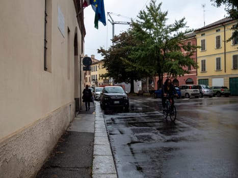 Cremona, Lombardy, Italy October 15th 2024 A cyclist is riding on a wet street in a small italian town, passing by parked cars and pedestrians