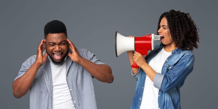 Communication problems. Angry black woman shouting with loudspeaker at her irritated boyfriend, standing together on light background