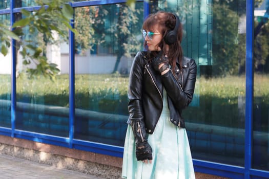 Image of a girl in a blue dress, black leather jacket, wearing blue glasses looking away against a blue glass wall. Adult Girl looking away.