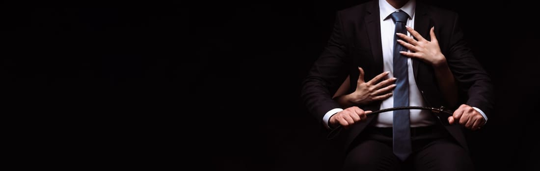businessman man in a dominant suit with a leather whip is sitting in a chair. Submissive girl puts her arms around him. BDSM concept. Panoramic wide horizontal photo for banner head cover
