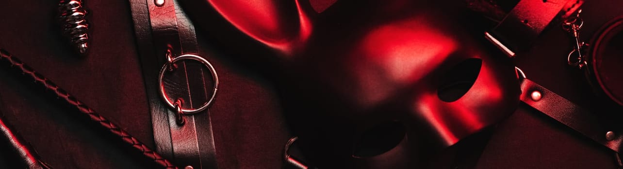 Set of erotic toys from a sex shop on a black and red background. Mask, leather belt, choker, handcuffs, whip and anal plug. Panoramic wide horizontal photo for banner head cover