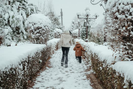 Happy cheerful mom with a child boy and son running backs in the snow in winter in the park