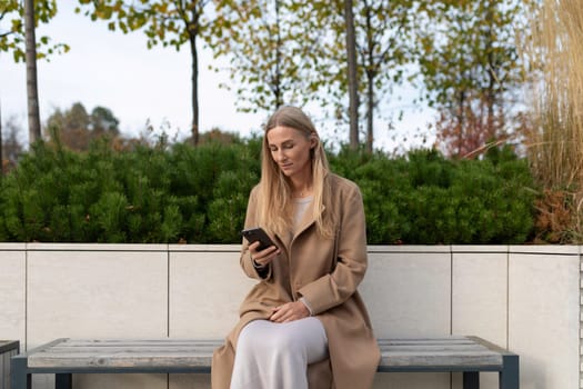 A woman with long blonde hair, wearing a long brown coat, is sitting on a bench. She is looking at a smartphone in her hands.