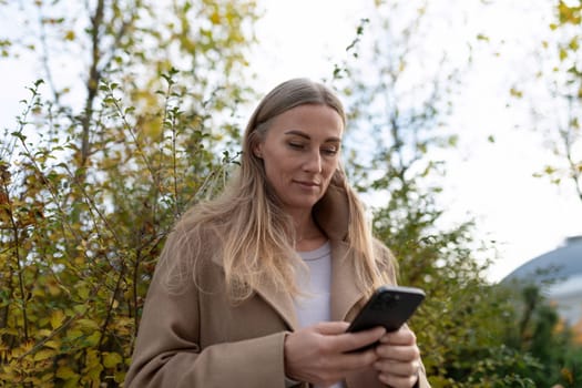 A woman in a brown coat stands near a building and looks at her phone. There are trees in the background.