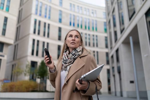 A woman is walking outside of a business center, holding a laptop and phone. She is dressed in a tan coat, scarf, and white shirt.
