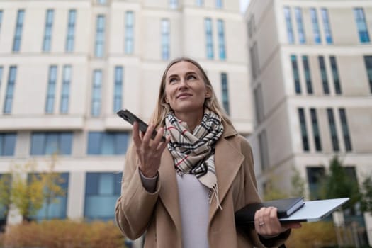 A woman in a beige coat and a scarf uses her phone while standing in front of an office building. She is holding a notebook and a laptop in her other hand, suggesting she's on her way to a meeting.