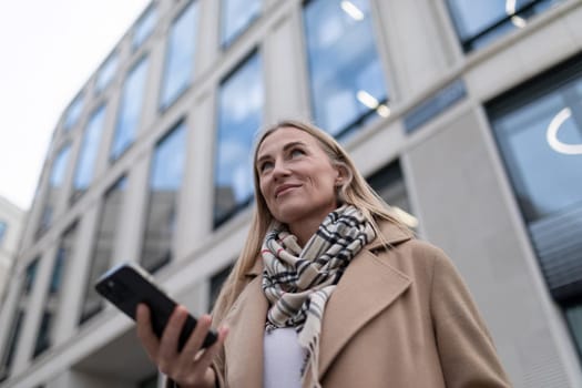 A woman wearing a long coat and a scarf is walking past a tall, modern building with many large windows. She is looking up and holding a black smartphone in her hand.