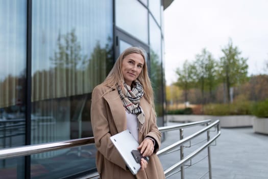 A woman in a tan coat and scarf is standing near a glass building, holding a laptop in her left hand. She is looking at the camera with a confident expression.