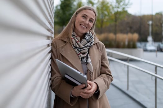 A woman in a brown coat and a plaid scarf stands leaning against a wall outside of a business center. She is holding a tablet in her hands. She is looking at the camera with a warm smile on her face.