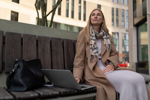 A woman wearing a tan coat and a scarf is seated on a wooden bench outside a building. A black bag rests next to her on the bench. She is using a laptop and looking up at something out of frame.