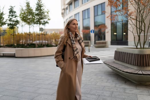 A professional woman strolls through a modern business district, carrying a notebook and appearing poised and determined amidst the autumn scenery.