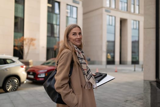 A business woman with long hair, dressed in a stylish coat, strolls confidently past a contemporary business center while holding a clipboard.